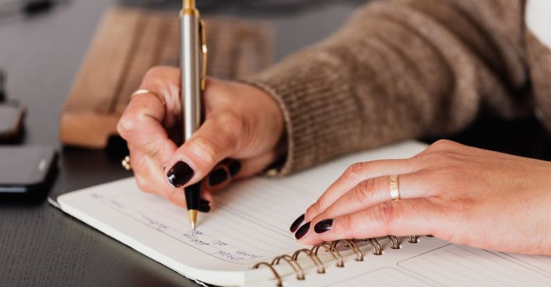 Business Plan Essentials - Crop unrecognizable female with stylish manicure sitting at black desk with keyboard and smartphone and taking notes with silver pen in notepad